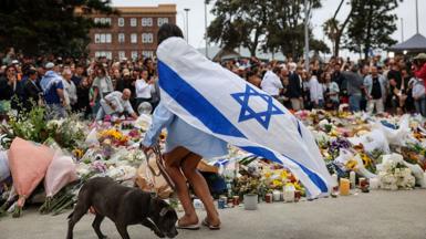A woman wearing an Israeli flag walks a dog in front of a floral memorial 