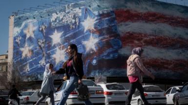 Young Iranian women walk past a state building covered with a giant anti-U.S. billboard depicting a symbolic image of the USS Abraham Lincoln aircraft carrier in downtown Tehran