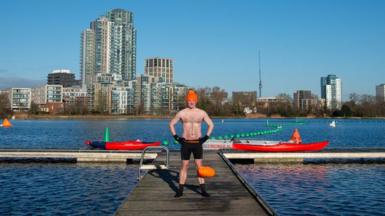 James stands on a ramp out across the west reservoir. He is wearing an orange bobble hat and swimming shorts as well as swimming socks. The sky is blue and there are high rise buildings in the distance, beyond the water's edge.