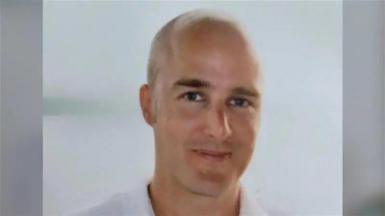 A clean-shaven bald man. He is smiling. He is wearing a white shirt against a white background. 