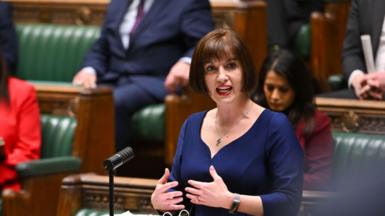 Bridget Phillipson speaks at a dispatch box in the House of Commons, with notes and microphones in front of her. Other attendees sit on the green benches, observing the proceedings.