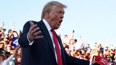 Donald Trump with his hands apart, holding a Make America Great Again cap in front of crowds at a rally