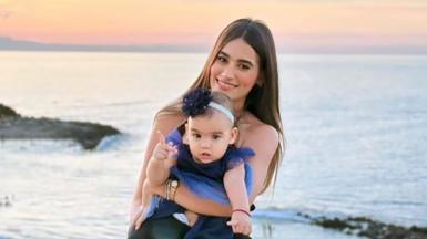 A woman with long dark hair smiles as she holds a toddler in a dress on a beach, the sunset behind her