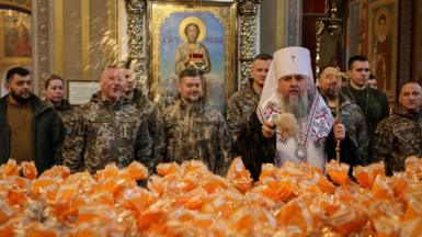 Metropolitan Epiphanius I, head of the Orthodox Church of Ukraine, sprinkles holy water on packets holding Easter cakes. Ukrainian service men stand behind him. 