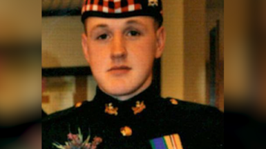 A young soldier in a Black Watch uniform and cap with red, white and black trim. He has medals and a flower on his uniform.