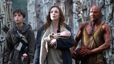 Alfie Williams, Jodie Comer & Ralph Fiennes in a film still from 28 Years Later. They are all looking up and in to the distance at the same thing off camera with worried facial expressions. Jodie Comer is in the centre of the trio and is carrying a baby 