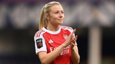 Leah Williamson, in a red Arsenal shirt with white sleeves and blonde hair tied back in a ponytail, looks in the direction of the crowd and applauds