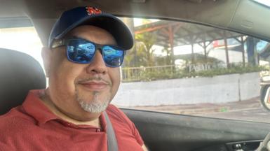 A man in the drivers seat of a car, wearing a blue cap and blue sun glasses looking into the camera.