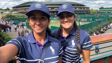 Aashny (left) and Saran (right) are seen above the tennis courts at Wimbledon. Both are wearing blue uniforms and caps as they are stood in the sunshine.