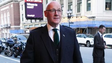 John Swinney, who is bald and wearing glasses, walks on a city street in formal wear 