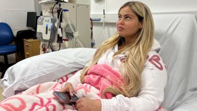 Kenzi Quinn sits in the Renal Unit at Sunderland Royal. She is sitting up in a bed and connected to a dialysis machine. Kenzi has blonde hair and is wearing a white hooded jumper while wrapped in a pink and white blanket, while holding her iPhone in her right hand. 