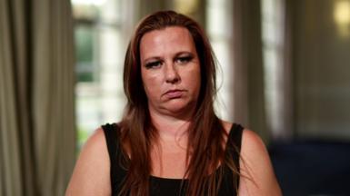 A woman wearing a black tank top and long reddish brown hair sits and stares at the camera.