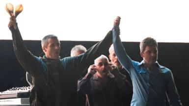 Liam and Noel hold their hands up in the air on stage 