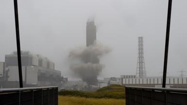Smoke surrounds the a tall chimney as it is demolished. It is a grey, foggy day with a white sky behind the chimney. A building can be seen to its left-hand side. 