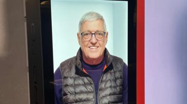 Hologram version of a man called Bill on a large screen, wearing a purple jumper and black gilet and smiling