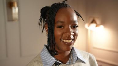 Corinna Brown is seen smiling widely in a portrait photograph. She is facing the camera and is wearing a grey jumper over a blue striped shirt