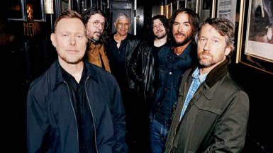 Six members of the Foo Fighters band stand side by side looking directly at the camera, hands in pockets.