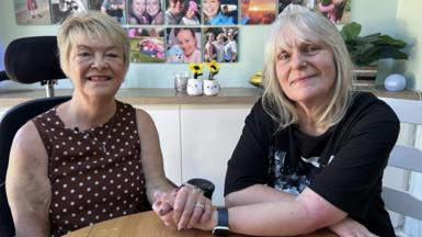 A woman with brown and white dotty top and short blonde hair sits in a wheelchair, hand in hand with a woman with shoulder-length blonde hair and black t-shirt. They both smile at the camera in front of a wall of photo tiles.