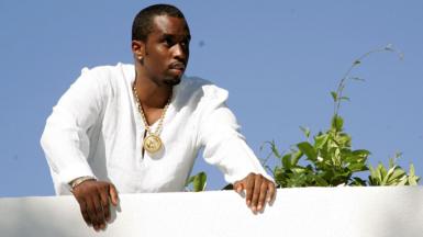 Sean "Diddy" Combs looks out over a white balcony wall while wearing a white top and gold chain, with a plant next to him and blue sky in the background, at one of his famous White Parties at his East Hapton estate in New York in 2007