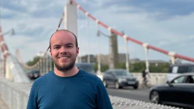 Young man in blue t-shirt standing in front of a suspension road bridge with cars in the background