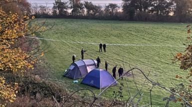 Two tents and a cordon on a greenfield with trees and woodland surrounding it. There are eight police officers in uniform standing near the tents