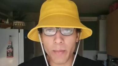 Nicholas Prosper, wearing glasses and a yellow bucket hat with a white string, talking into the camera in a homemade video in a kitchen