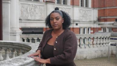 Nadine is standing outside, looking at the camera. She has black curly hair and is wearing a brown cardigan.