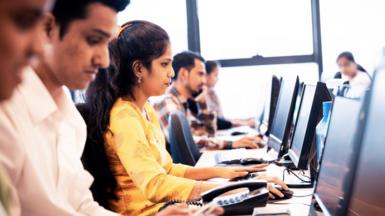 The image shows Indian workers at a software company seated in front of their computer screens. A woman wearing a white kurta is prominently seen, surrounded by male colleagues on either side. 