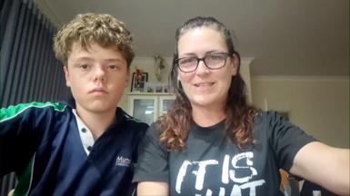 Austin Applebee, who has blonde curly hair and is wearing a navy top with green stripes down the arm, sits next to his mother Joanne, who wears glasses and has brown hair, in a living room