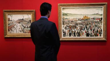 A man stands and admires two paintings by the artist LS Lowry, which depict an industrial landscape oif houses and factories and a fairground with a crowd of people