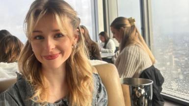 Eve Dulcie sits inside a in a rooftop restaurant with a skyline in the background. She smiles at the camera. She has blonde mid length hair, which is half pulled back from her face with her bangs loose in her face. 
