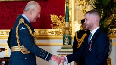 King Charles in RAF uniform smiles at Nathan Newby, a man in a dark blue suit, as he pins a medal on him. In the background is another man in military uniform who stands in a grand-looking room with gold decorations and a carved marble fireplace.