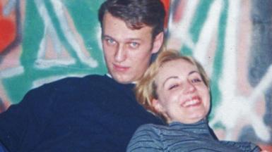 Alexei Navalny and his wife Yulia in their youth, the photo is cropped tight showing their head and shoulders as they embrace in front of a graffiti'ed wall