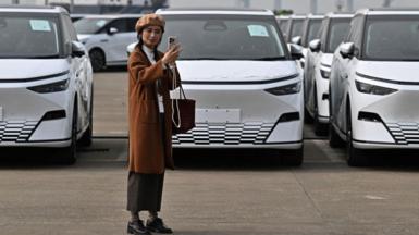 A woman takes a selfie in front X9 electric vehicles by Chinese EV manufacturer XPeng, are loaded on ship of the NYK line during a ceremony in the Port of Guangzhou, China's southern Guangdong province, on 22 February, 2025.