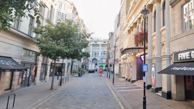 A pedestrian‑friendly cobbled street in central London with shops, trees, and a Five Guys restaurant on the right.