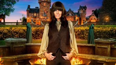 Claudia Winkleman stands in front of a firepit and behind that the Traitors castle in the distance and smiles at the camera. Two hooded Traitors are visible at either side of Claudia in the background