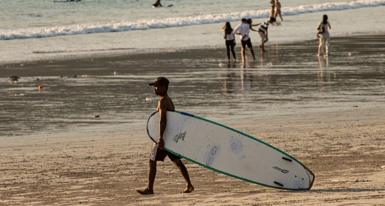 A man dragging his surfboard towards the sea on a beach. He is wearing board shorts and barefoot, walking on the sand. In the background are some people walking near the shoreline.