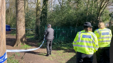 Two female police officers wearing yellow high-vis jackets watch a male officer, wearing black, stand by police tape. In the distance a blue police tent can be seen behind trees in the park