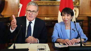 UK Prime Minister Sir Keir Starmer and Japanese Prime Minister Sanae Takaichi sitting a table in 10 Downing Street in London. Starmer is on the left wearing a dark suit, white suit, and navy blue tie with white dots. Takaichi is on the right wearing a blue jacket, blue top, and a white necklace. Behind them, there is a UK flag and a Japanese flag. Behind that, there wood-panelled wall and the bottom of an ornate frame of a painting is just visible.