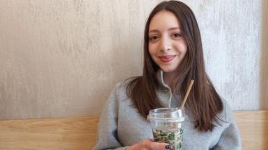 A woman with long brown hair in a grey coat smiles at the camera as she holds up a brown drink in a plastic cup with a straw. She is sat in front of a plain grey painted wall with a wooden plank.