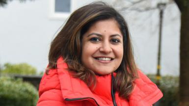 A woman with shoulder-length brown hair smiles. She is wearing a large salmon-coloured puffer jacket.