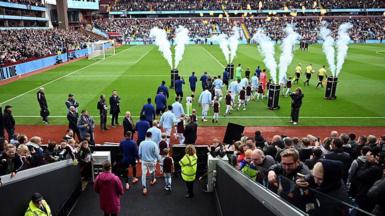 Players in light blue uniforms walk out onto the pitch at Villa Park, as smoke flares up and a packed crowd watches from the sidelines.