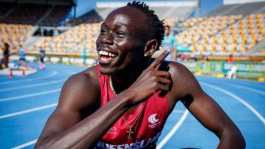 Gout Gout, wearing a red vest and a gold crucifix around his neck, points with his right hand across his body and smiles while crouching on a blue athletics track with white and yellow stadium seating in the background. 