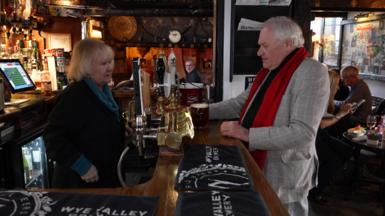 Two people in a pub. The woman behind the bar has blonde hair and a black top with turqouise top underneath. She is talking to a man with grey hair, a grey jacket and red tie