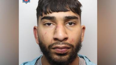 A mugshot of an Asian man in his early 20s, who has black hair cut into a fringe and black facial hair. He has red marks to his face and is looking into the camera with a blank expression. 