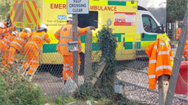An ambulance is surrounded by people in high vis jackets pushing it from the back. It is sitting on gravel and train tracks.