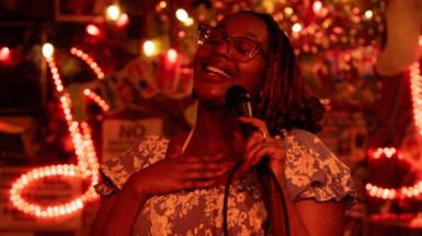 Edna Nicole Luckett sings at a microphone, her hand on her heart, with light-up signs showing musical notes behind her. She's wearing glasses and a floral dress, and her eyes are closed.