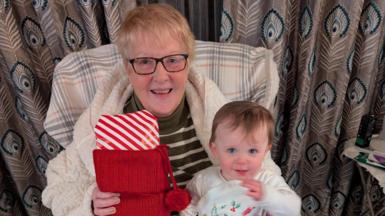 Grandmother Pat grant sitting on a chair smiling at camera holding her baby granddaughter and a Christmas stocking. They're both smiling. 