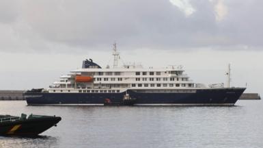 A cruise ship sits on the water, with a smaller vessel alongside, the front of another boat can be seen in the foreground to the left