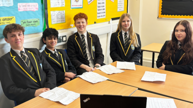 Five Year 10 students sit around two desks pushed together. They all wear a white shirt, a navy, white and gold striped tie, navy blazer with gold edging on the collar and a school badge on their left. Kit is white with short light brown hair. Jake has olive skin with short black hair. Hugo is whit with slightly curly red hair which is cropped at the sides. Cali is white with long blonde hair. Maisy is white with very long brown hair which is slightly wavy at the bottom. There are pinboards with phrases in Welsh behind them.